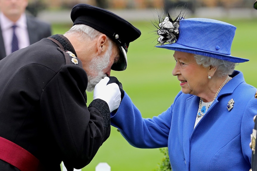 Un uomo in uniforme militare bacia la mano della regina che indossa un soprabito blu e un cappello abbinato. 