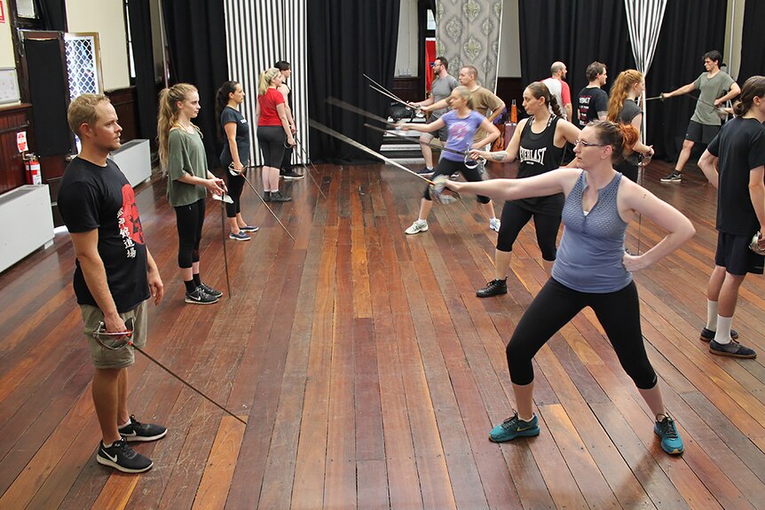 A group of actors stand in two rows with fake swords in their hands pointing at each other.