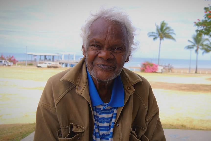 Aboriginal man with shirt and jacket looks at camera with beach background.