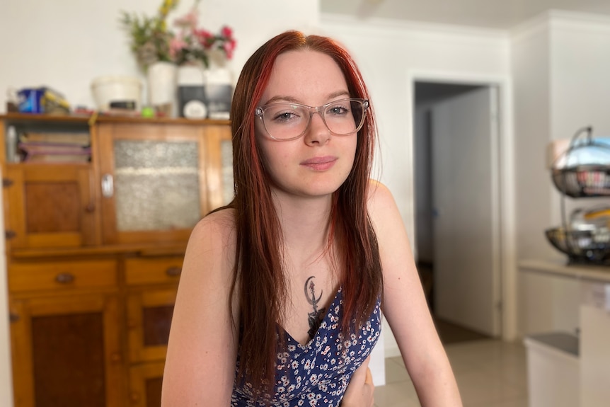 A young woman with long brown hair and glasses looks at the camera, she is holding her ribs