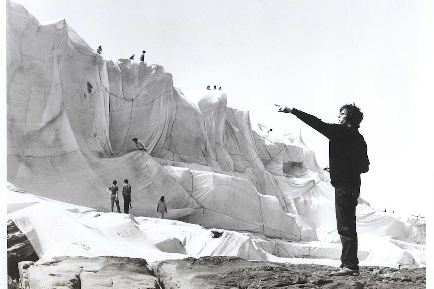the artist Christo stands in front of wrapped coast pointing to something in the distance.