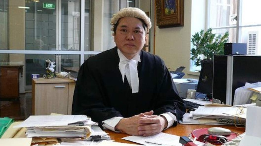 A man wearing a barrister's wig and robes sitting at a desk with papers on it