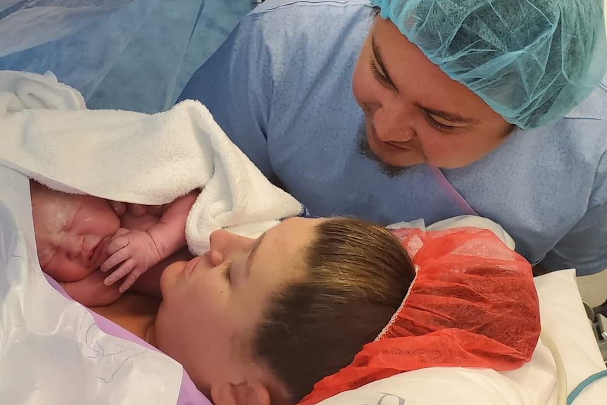 A woman with a red hair net lies in a hospital bed holding a newborn baby.