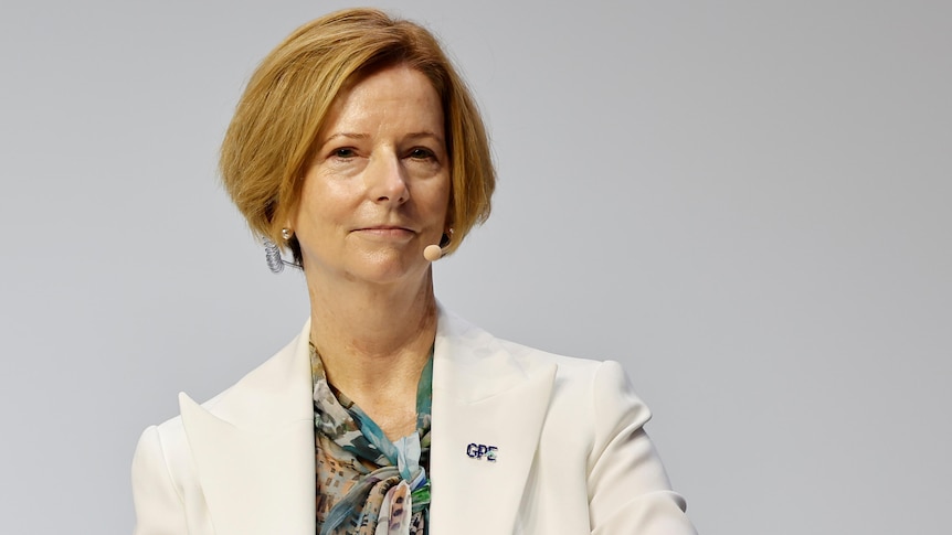 Julia Gillard from torso up sitting on stage for a talk holding sheets of paper. 