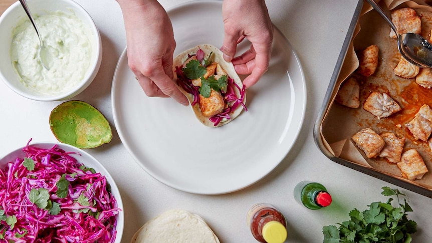 A fish taco sits on a plate with a bowl of avocado yoghurt sauce beside it and a bowl of cabbage slaw, an easy dinner for kids.