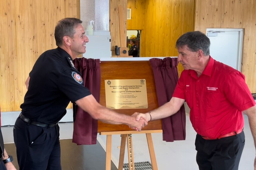 Two men, one in all black uniform, the other in a red t-shirt and black pants shake hands in front of an unveiled plaque.