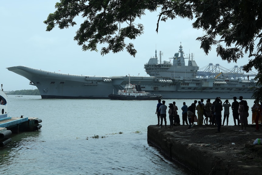 People in silhouette watch a large aircraft carrier move past land and out to sea.