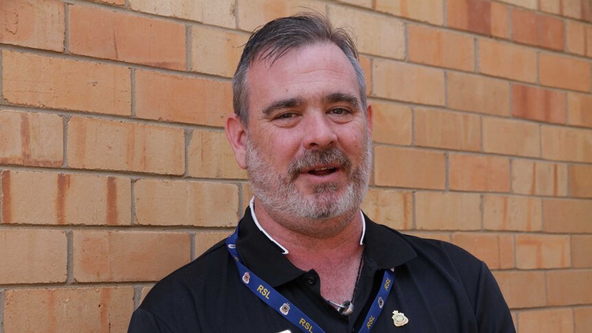 A bearded man wearing a dark shirt with a lanyard around his neck stands against a pale brick wall looking at the camera.