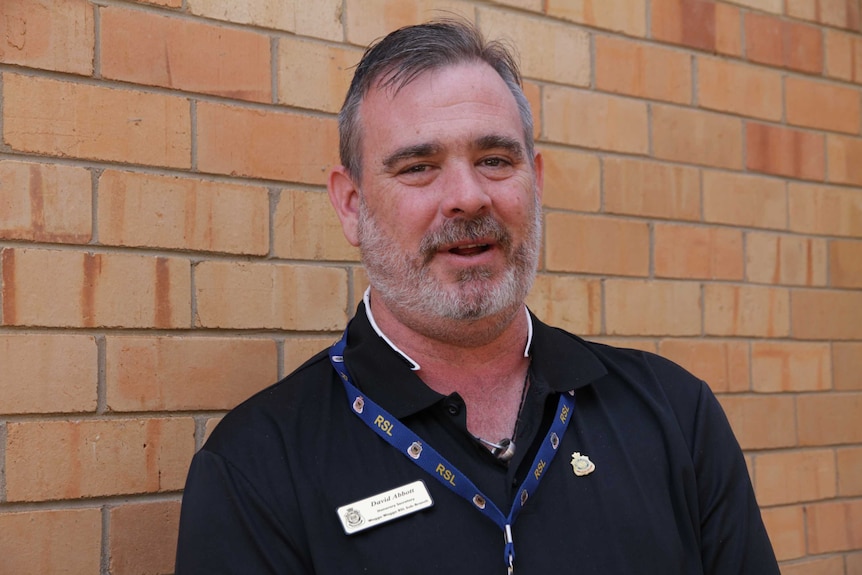 A bearded man wearing a dark shirt with a lanyard around his neck stands against a pale brick wall looking at the camera.