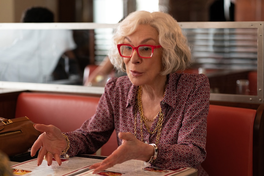 Jeannie Berlin, an older white woman with wavy white hair and red glasses, sits at a diner talking to someone off camera.