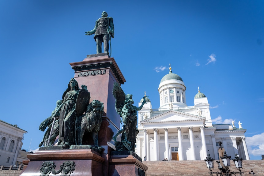 The statue of Alexander II stands in front of a historic white building in Helsinki.