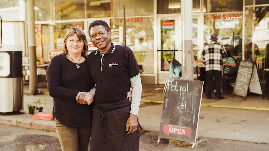 A woman in a black shirt smiles for a photo with her arm around a man, also smiling, in front of a shop window.