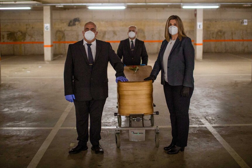 Three people in face masks surround a coffin in an empty car park