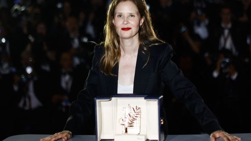 Director dressed in black jacket stands behind her Palme d'Or award, smiling. Crowd of photographers behind her. 