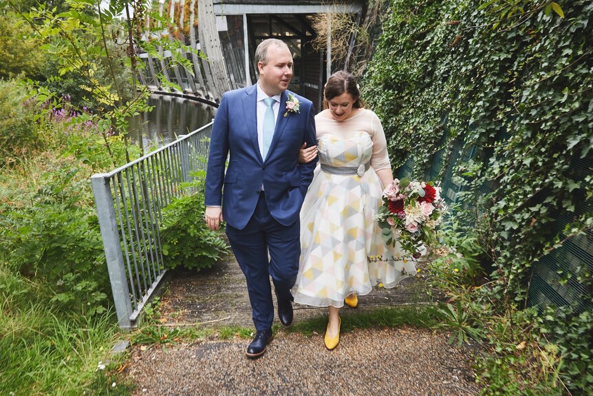 A couple standing in a garden, walking up stairs, arms clasped together, man in a suit, woman wearing white and yellow, flowers 