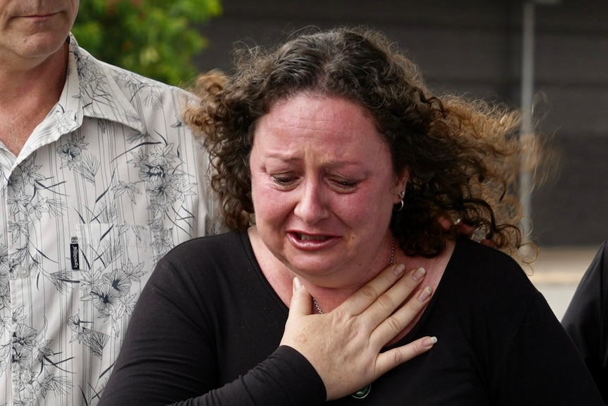 A middle-aged woman with curly windswept hair cries while her hand is on her chest.