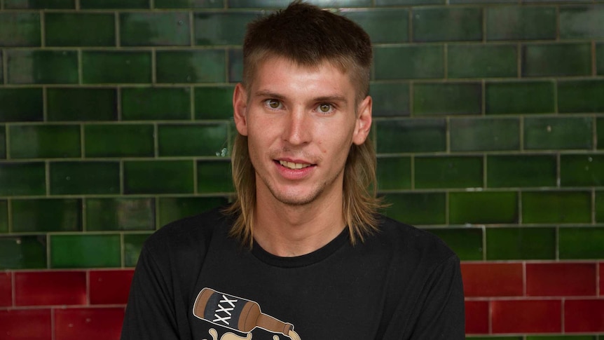Young man with a mullet hairstyle poses in front of a green title hotel wall.