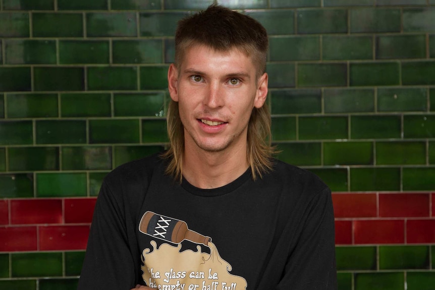 Young man with a mullet hairstyle poses in front of a green title hotel wall.