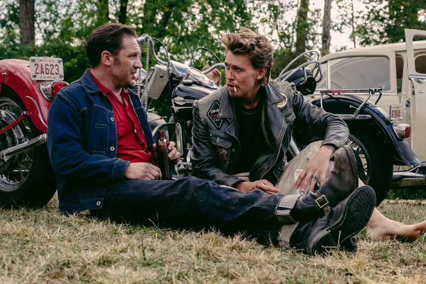 Tom Hardy sits on the ground next to Austin Butler, both in motorcycle leathers, drinking from bottles