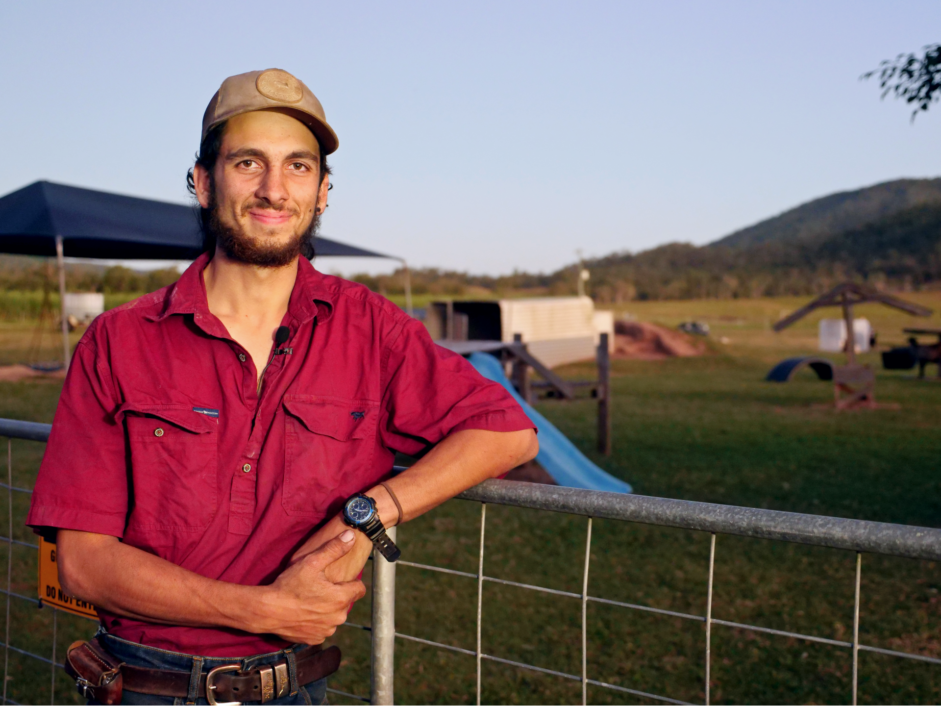 a young man in a farming shirt and cap leans against the fence of a paddock, smiling at the camera