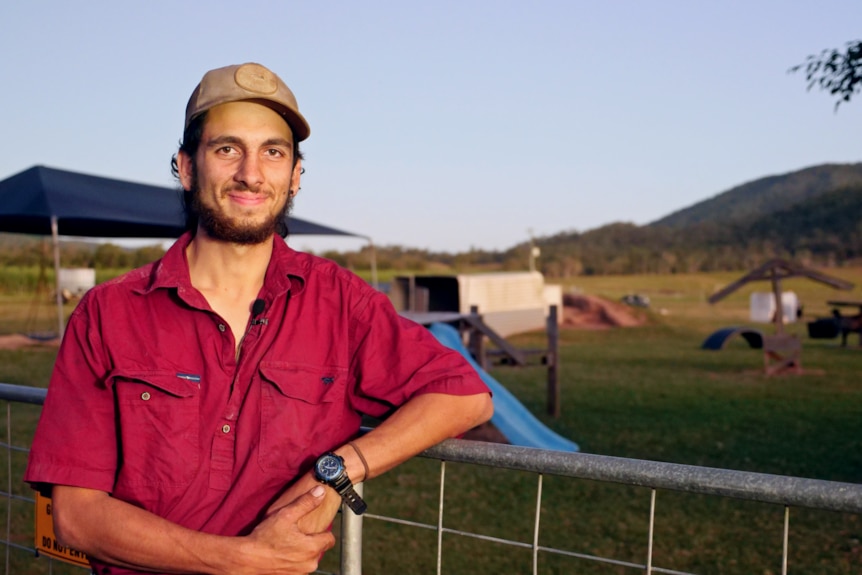 a young man in a farming shirt and cap leans against the fence of a paddock, smiling at the camera