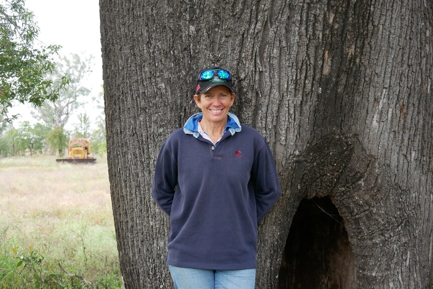 A smiling woman wearing a cap stands in front of a tree.
