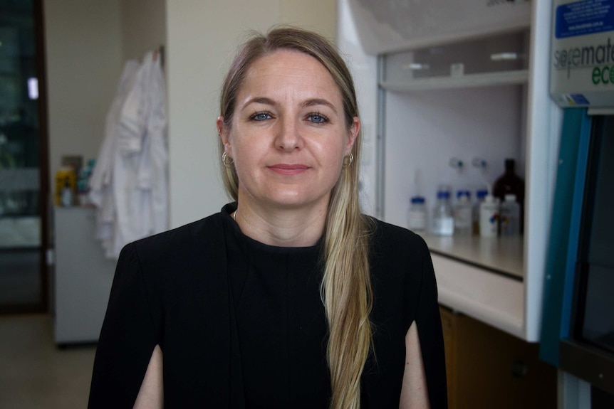 Image of a woman with blonde hair, she's wearing a dark shirt and standing in a laboratory.