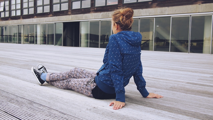 A young woman wearing a blue hoodie, tights and sneakers sits on a concrete floor outside a building