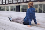 A young woman wearing a blue hoodie, tights and sneakers sits on a concrete floor outside a building
