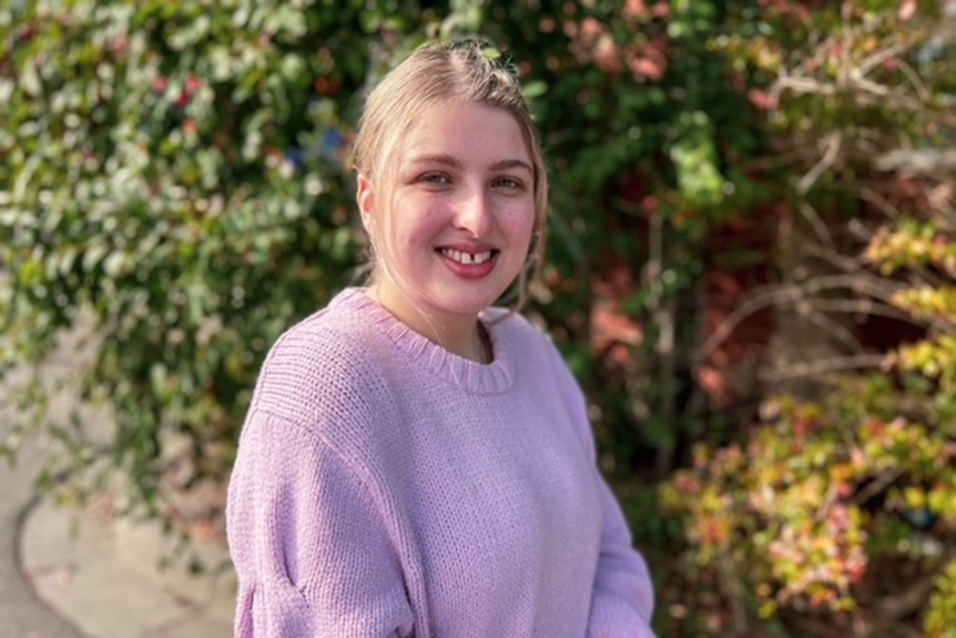 a blonde woman in a purple jumper smiles at the camera. there are plants behind her.