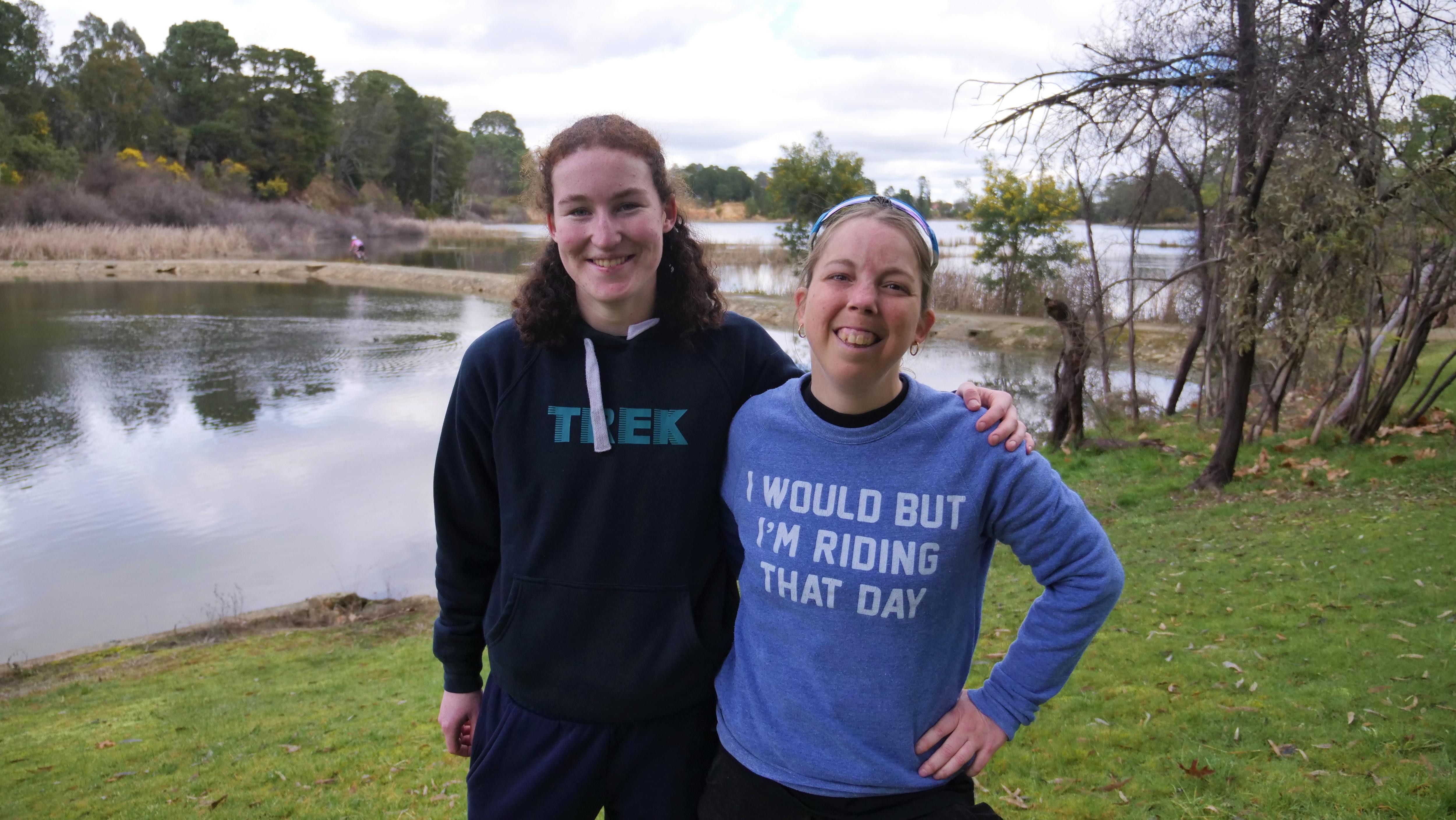 Two women stand in front of the lake smiling.  One is wearing a jumper that says: I would but I'm riding that day. 