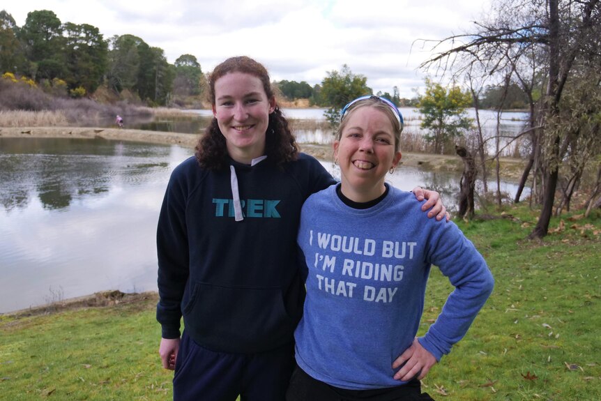 Two women stand in front of the lake smiling.  One is wearing a jumper that says: I would but I'm riding that day. 