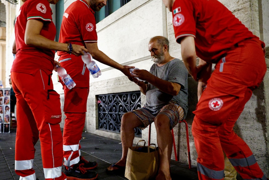 Three Red Cross aid workers give water to a homeless man sitting on a chair outside.
