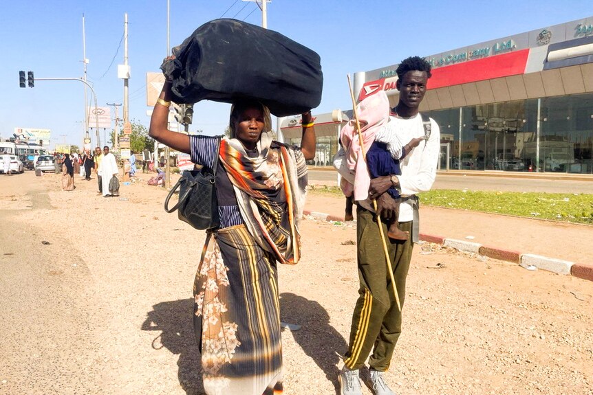 A woman carries a suitcase on her head while a man walks beside her carrying a baby.
