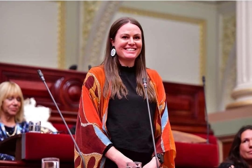 A woman, with long brown hair, stands and smiles