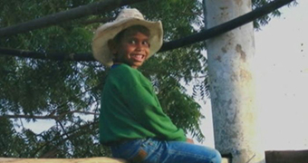 A young Aboriginal boy smiles while wearing a cowboy hat and sitting on a log fence.