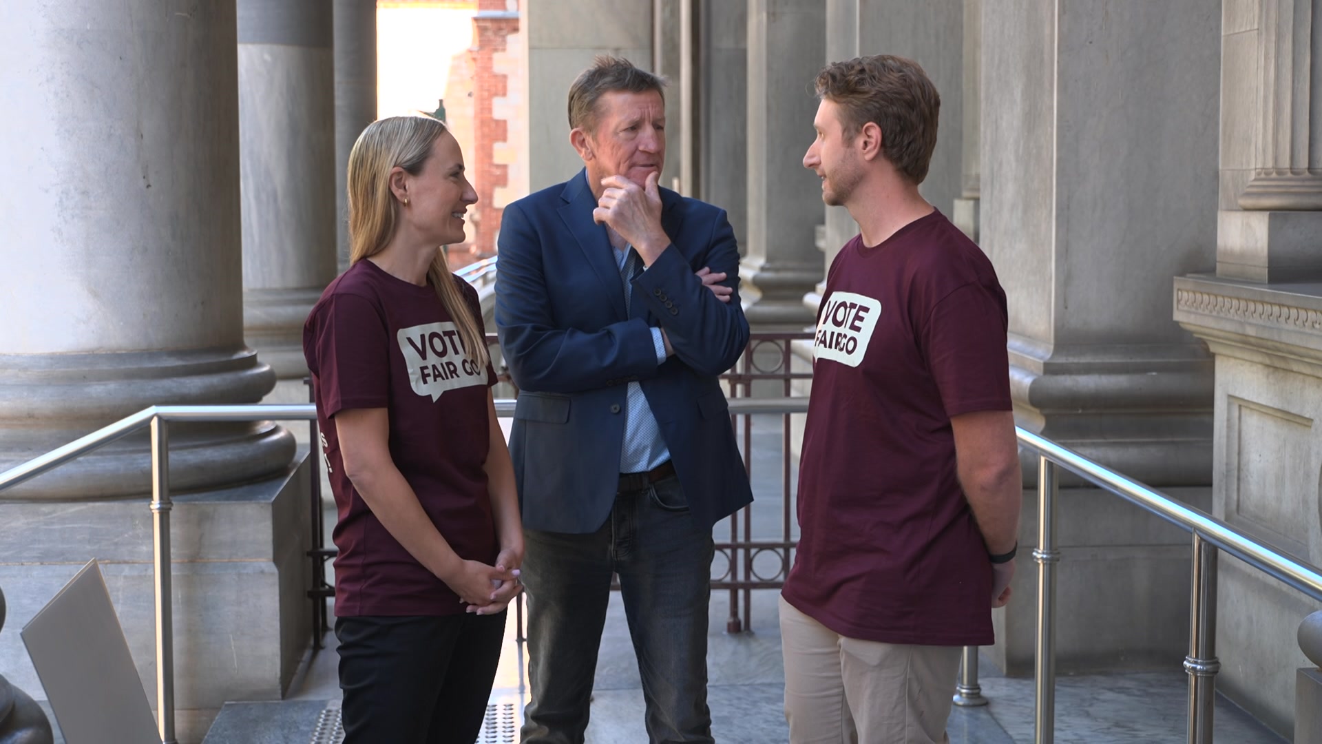 A woman wearing a maroon T-shirt saying VOTE FAIR GO speaks with a man wearing a suit and a man wearing the same as her