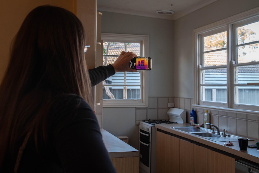 A woman holds a phone with thermal imaging app open up inside a kitchen with bright light coming from windows