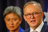 Anthony Albanese, with Penny Wong behind him, answes questions in front of an australian flag