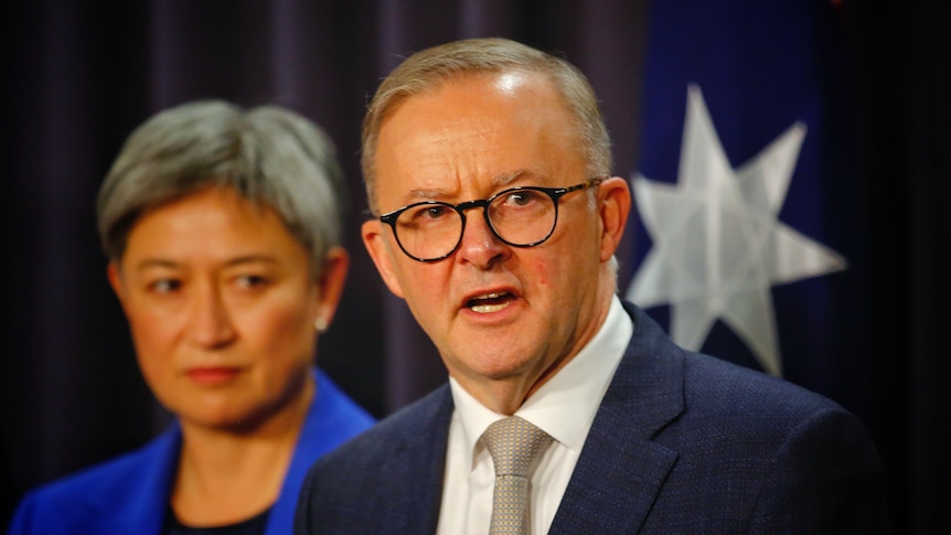 Anthony Albanese, with Penny Wong behind him, answes questions in front of an australian flag