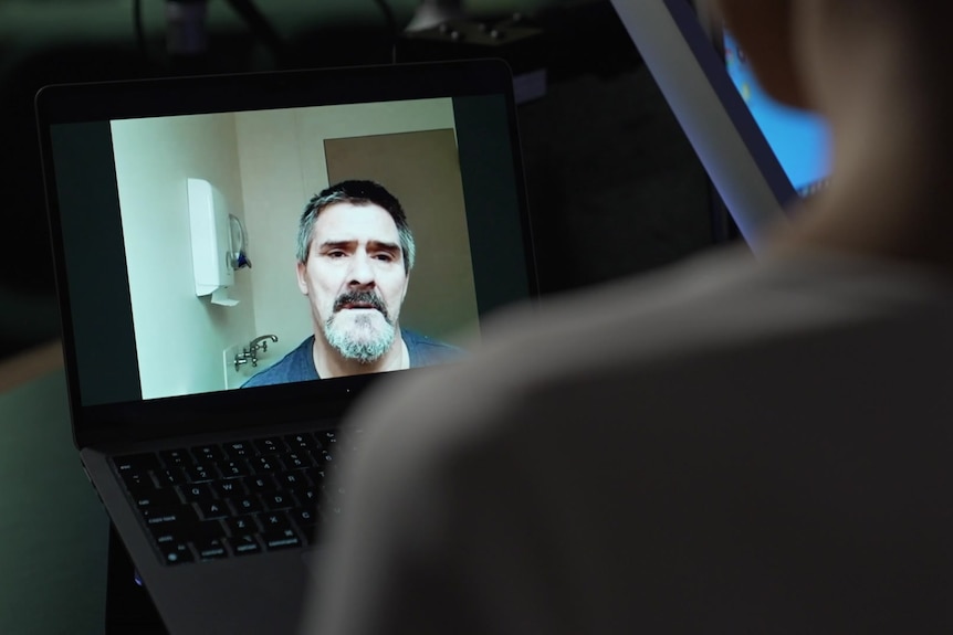 A man on a laptop screen speaks to a reporter who is pictured from behind