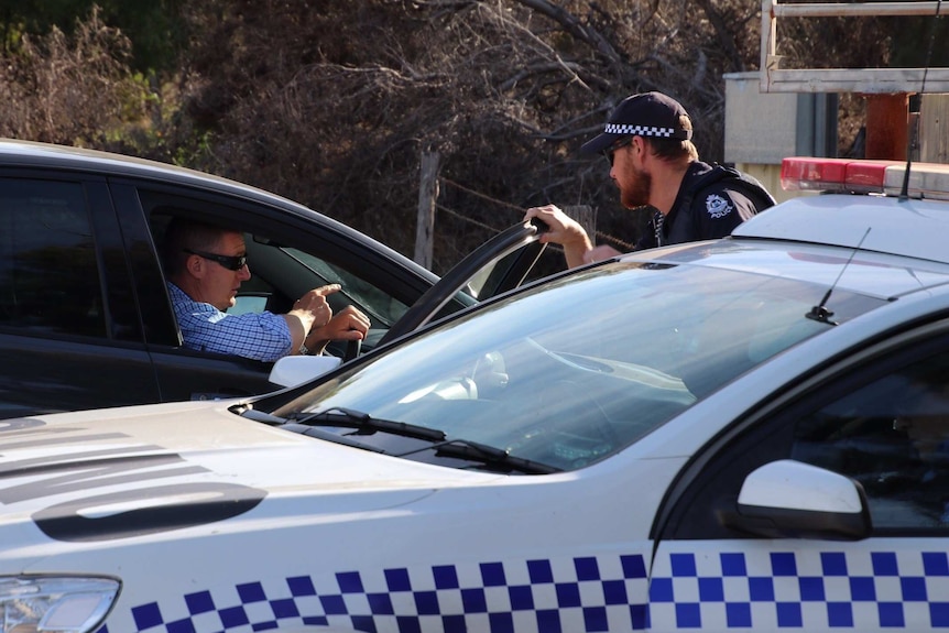 An officer in a police car speaks to a man in a black car wearing sunglasses.