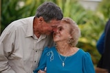 An elderly married couple warmly embrace while smiling.
