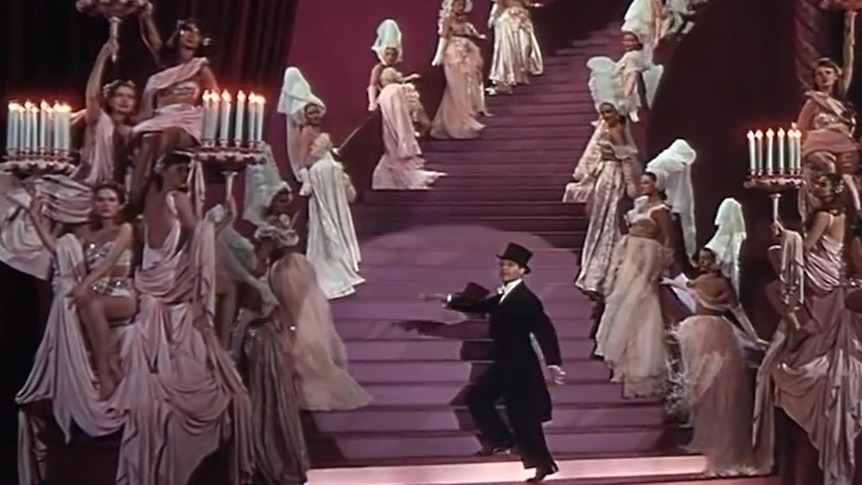 A man stands on a pink staircase in a suit and top hat, surrounded by women in pink holding ornate candelabras.