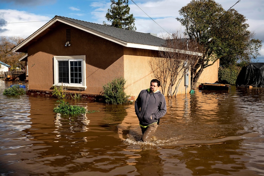 A man in a grey jumper knee deep in flood water outside a house