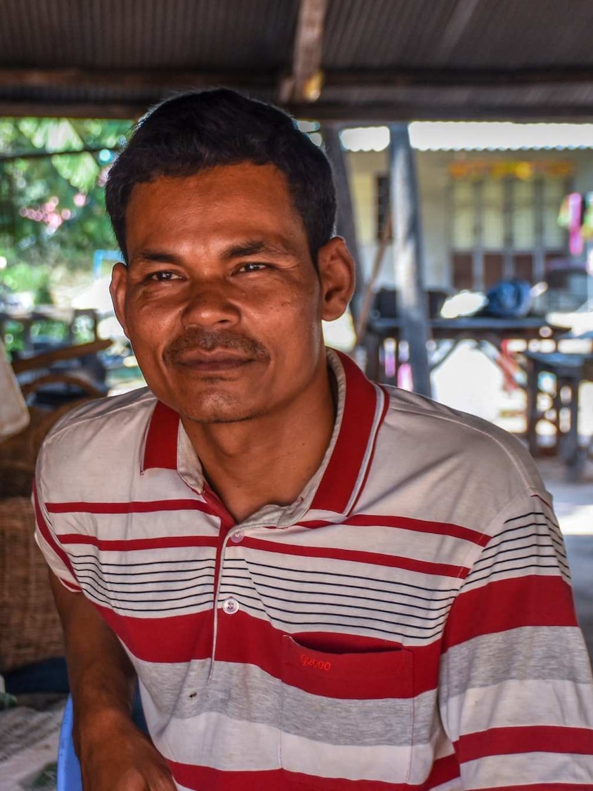 A smiling man sits outside his wooden house