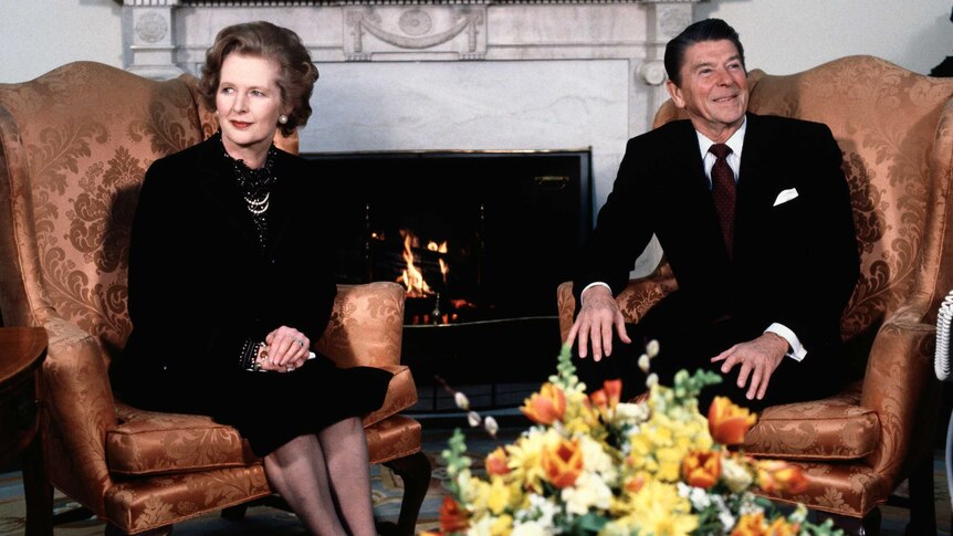 Margaret Thatcher dressed in black sits with Ronald Reagan, who is dressed in a dark suit, by a fireplace.