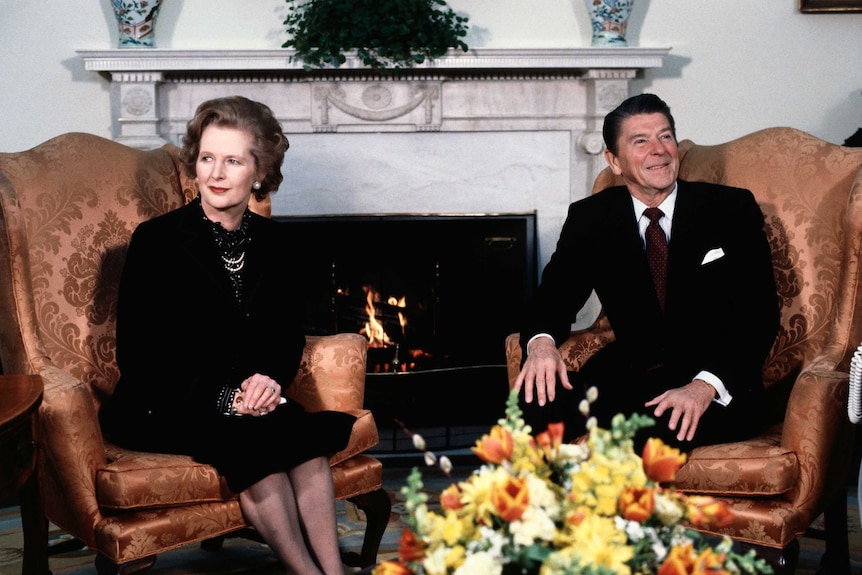 Margaret Thatcher dressed in black sits with Ronald Reagan, who is dressed in a dark suit, by a fireplace.