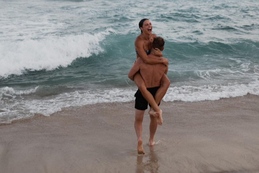 Luke is wearing swimming shorts and walks towards the ocean. He is carrying a woman in his arms. She is laughing.