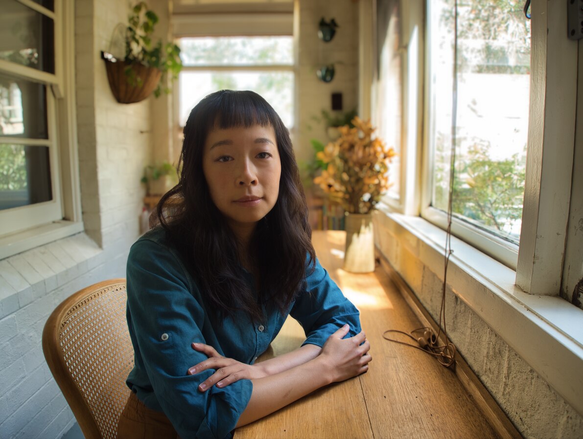 An Asian Australian woman sits at a desk in sunlight, her arms folded in front of her
